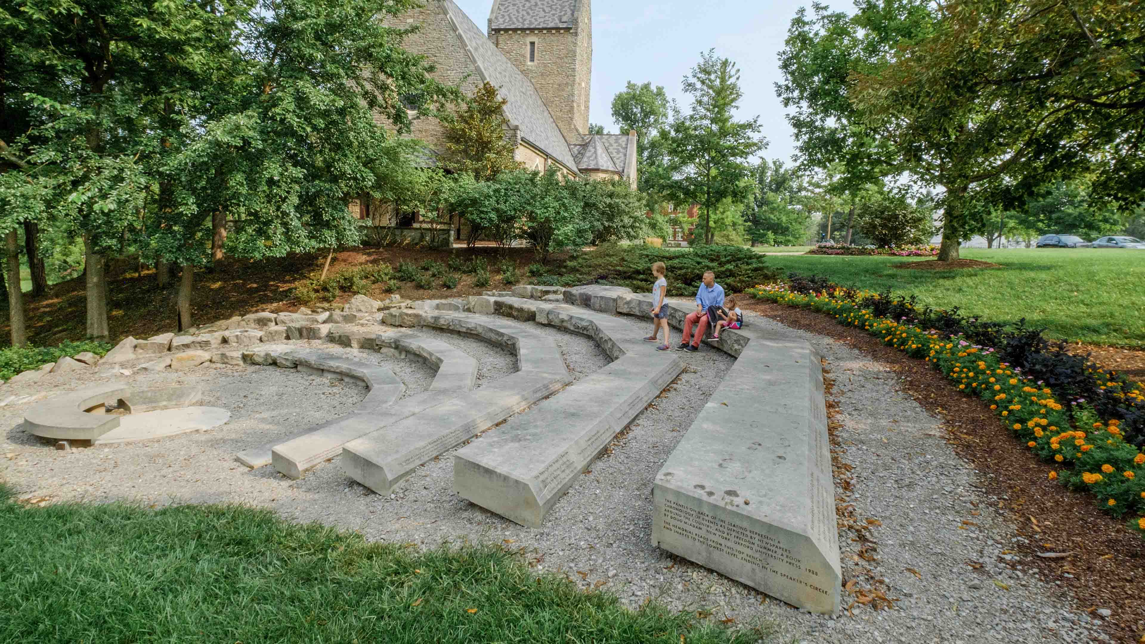 A man and a young girl sit on a stone bench, which is part of the Freedom Summer Memorial on Western Campus of Miami University. A second young girl stands on an adjacent stone bench, and all three people look toward the center of the memorial.