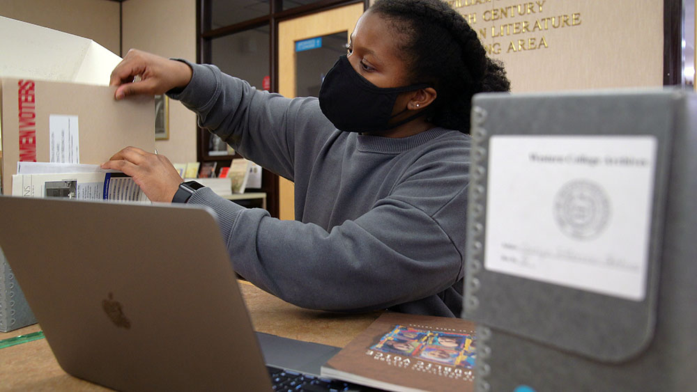 A young Black female student employee of the University Libraries wearing a face covering sits at a table in the Walter Havighurst Special Collections and Archives Reading Room with a laptop and archival storage boxes containing materials donated by Carolyn Jefferson-Jenkins. The student is taking a folder out of one of the boxes.