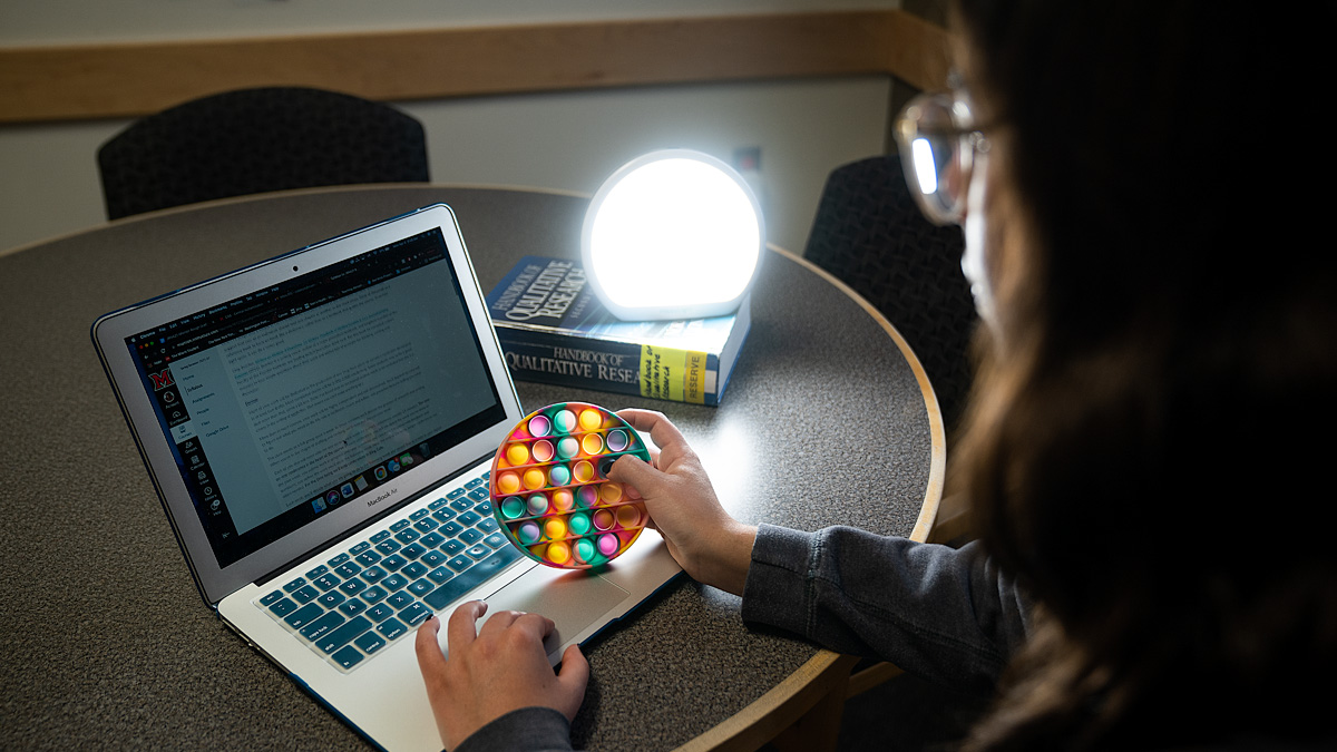 A student uses a bubble fidget while reading on a laptop. A full-spectrum LED lamp sitting on a textbook shines toward her.