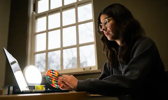 A student holds a bubble popping fidget in a library study room while using a laptop. A bright therapy light is shining on them from the table. A textbook is also on the table.