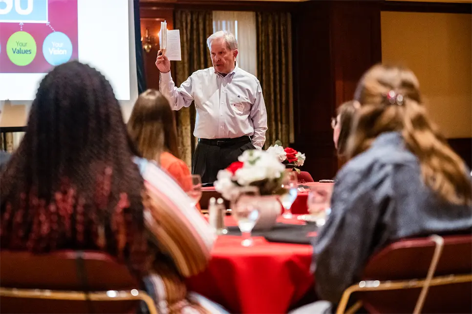 Tom Heuer holds up a book as he speaks to students seated at tables in the Shriver Center at Miami University. A projector screen behind him displays a slide with the words: Your Values and Your Vision.