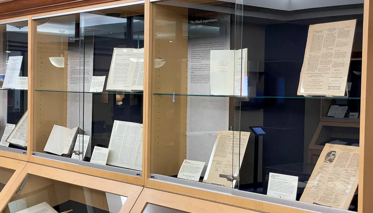 A photograph of display cases for the Poetry at Miami exhibition. Several books and pages are propped up in the display cases with small label cards adjacent to them. There are large panels in each of the cases behind the items with headers for the different themes and eras for the pieces in that case. The one closest to the camera reads: The Black experience, 1960-1980
