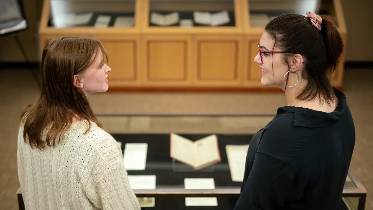 Two students look at one another and talk inside the exhibition room on the third floor of King Library. A glass display case stands in front of them in the center of the room containing documents and open books. Another display case, against the wall further on, contains more documents and open books.