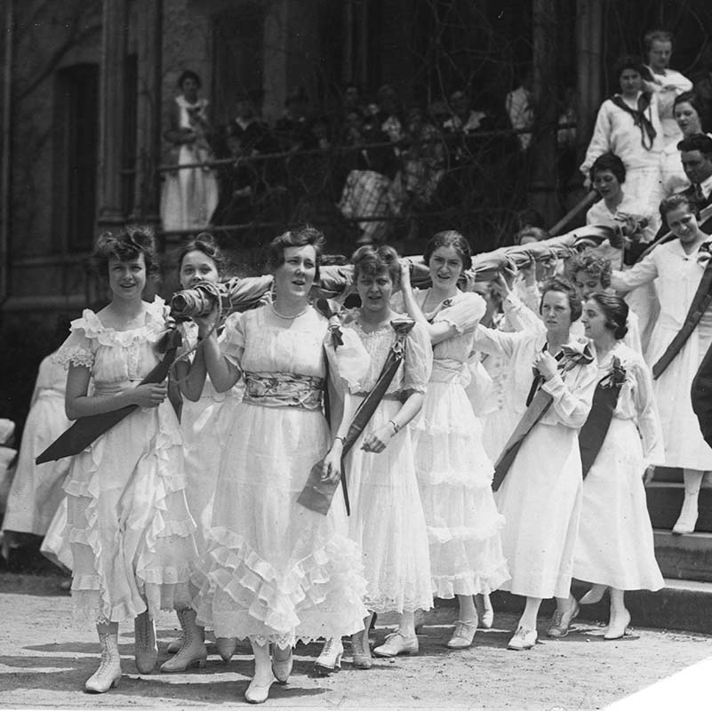 Group of female Western College students in long white dresses carry an unfurled oversized flag